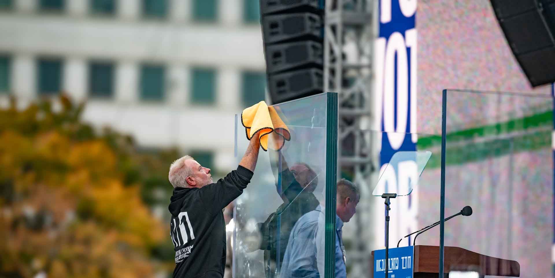 PM1 crew member wiping down the glass panels around the podium at an outdoor rally