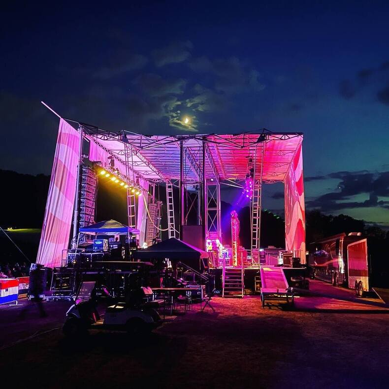 Outdoor PM1 stage lit pink and purple with scaffold towers and a canopy