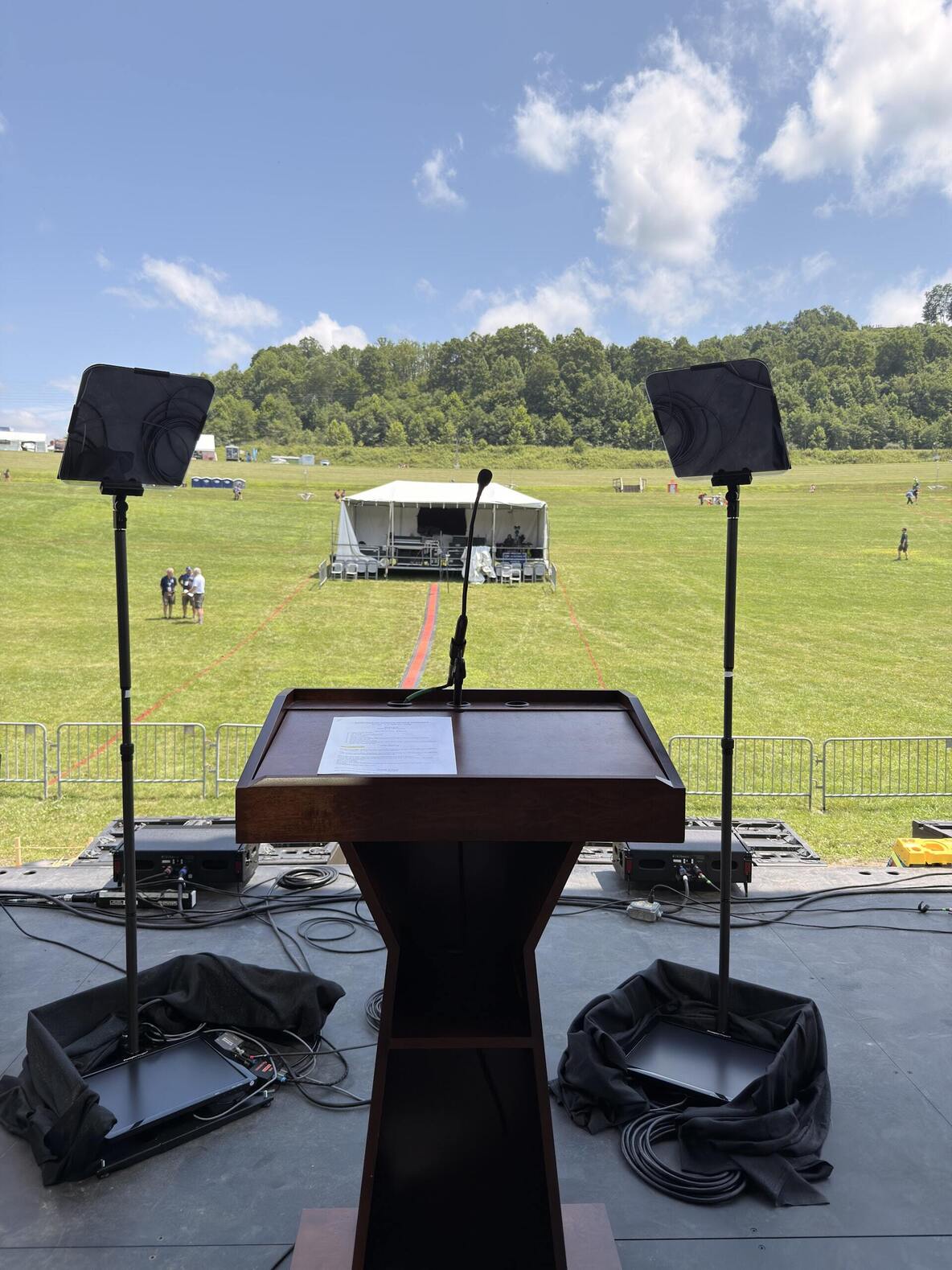 View from behind a podium at the 2026 Scouting America Jamboree and teleprompter glass on an outdoor stage, looking out over a field