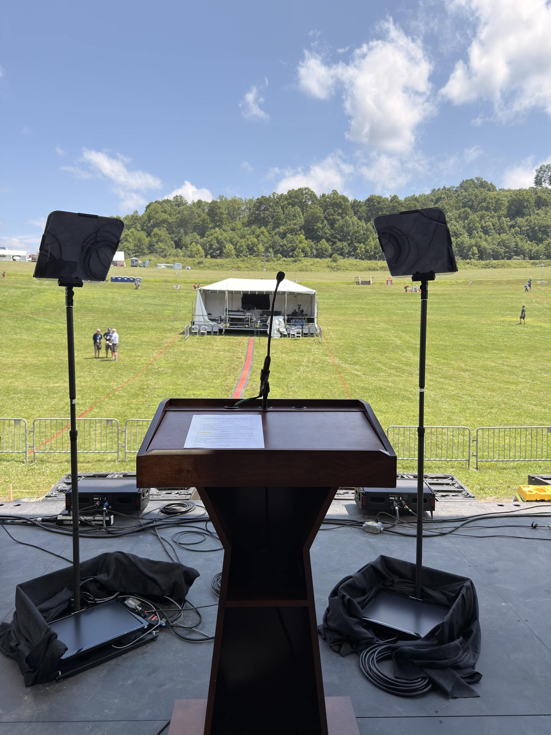 View from behind a podium at the 2026 Scouting America Jamboree and teleprompter glass on an outdoor stage, looking out over a field